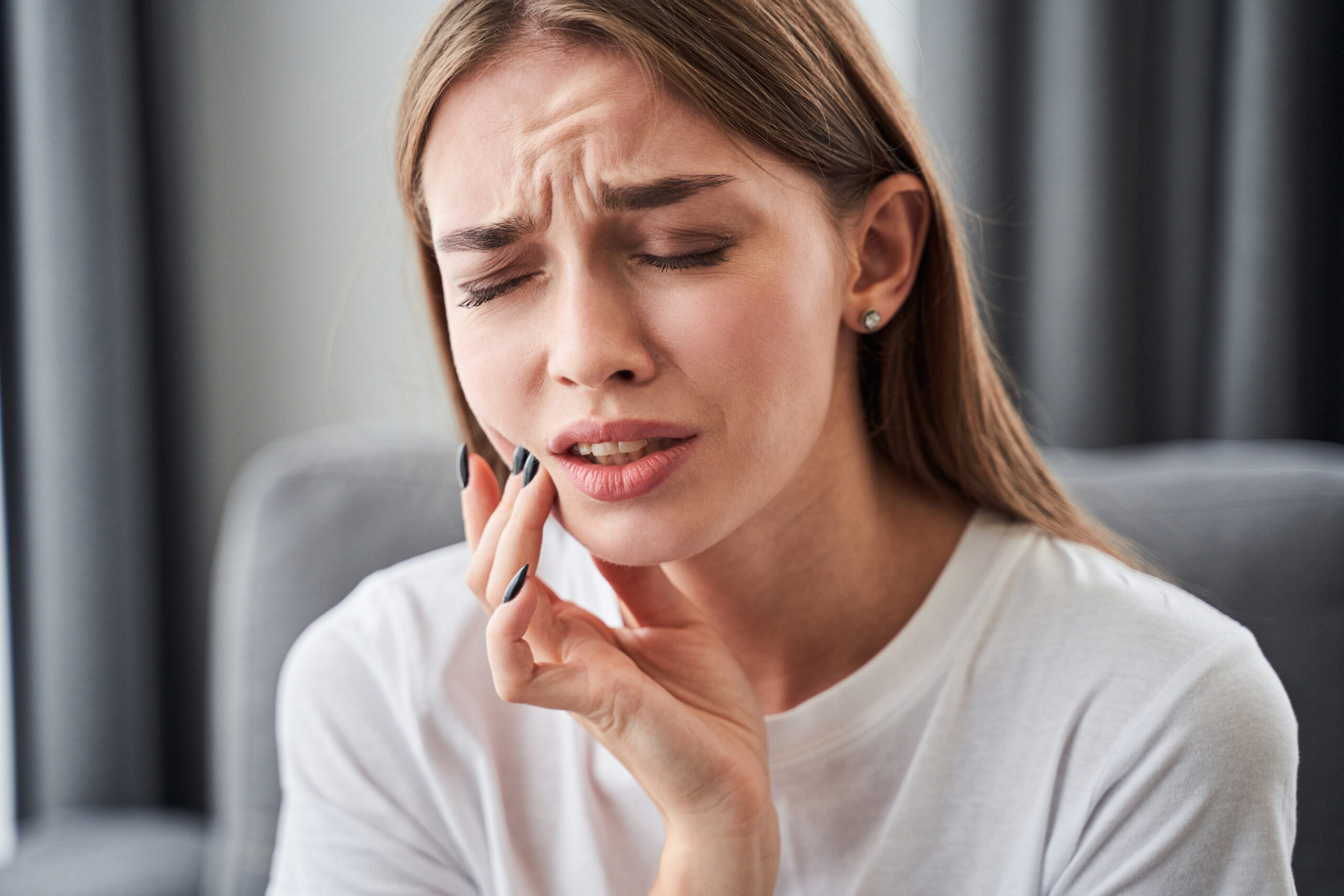 Girl touching her cheek with her fingers Can not take the pain. Frustrated young women suffering from toothache and touching her cheek with her fingers while sitting on the sofa at home